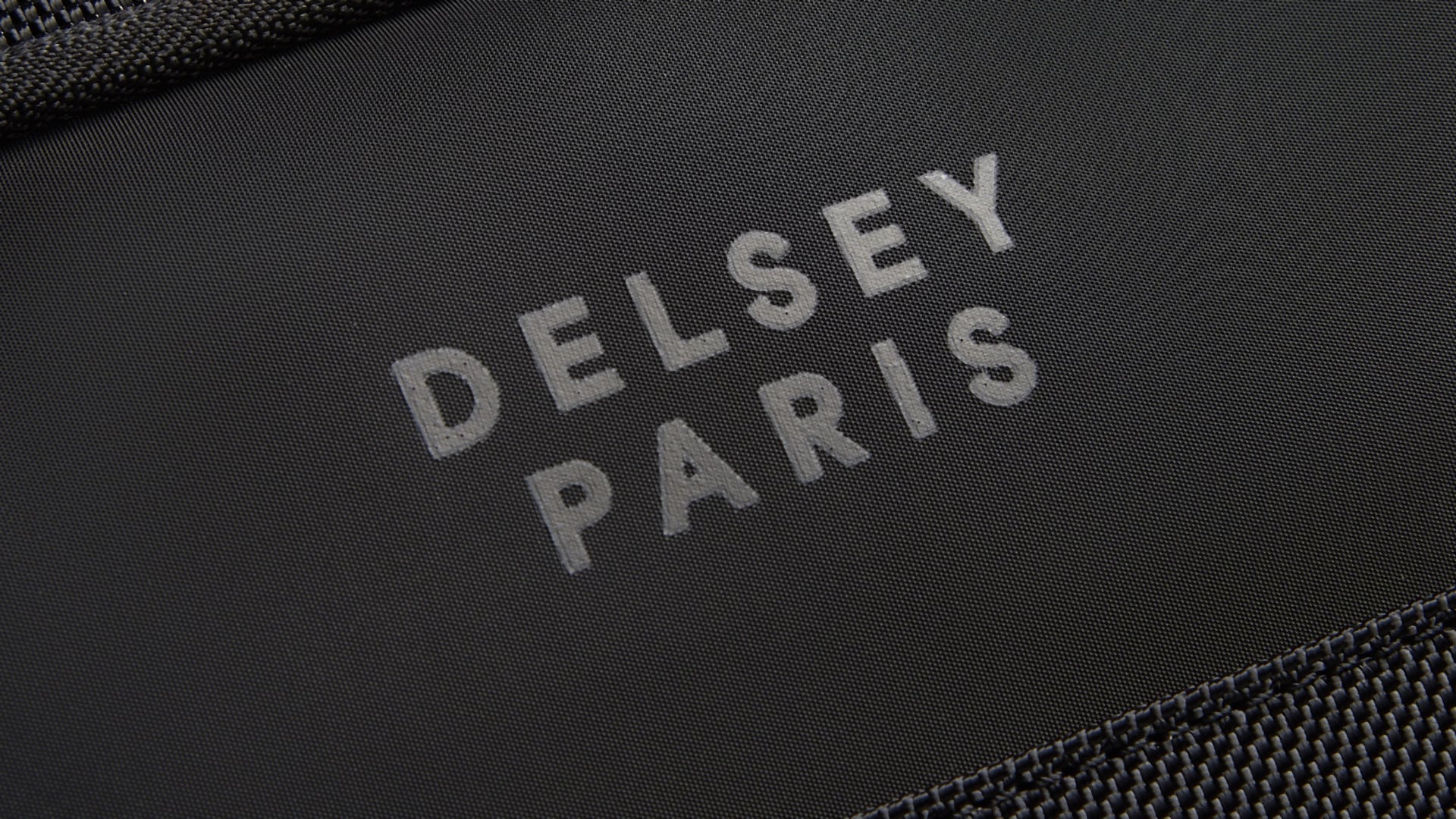 Gros plan sur le logo DELSEY PARIS imprimé sur le panneau avant d’une valise business Parvis Plus noire, entre deux bandes de tissu texturé